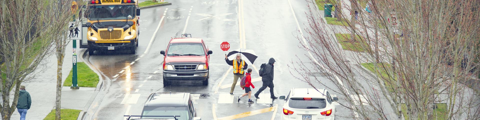 A crossing guard helping children cross a busy road with buses and cars in the rain.